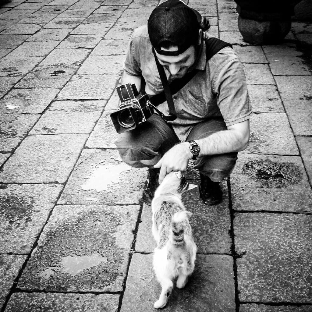 David is all about cats and cameras. A monochrome photograph of David R Munson holding a Hasselblad camera and petting a cat at a temple in China